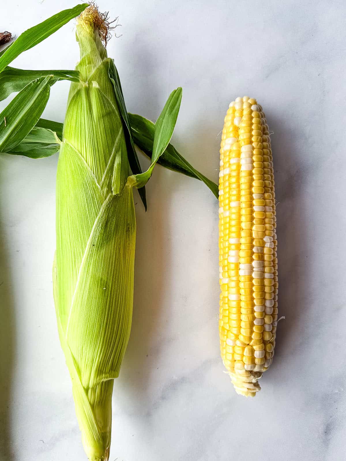 Two fresh ears of corn on the counter. The one on the left has a bright green hull. The one on the right has had the hull removed.