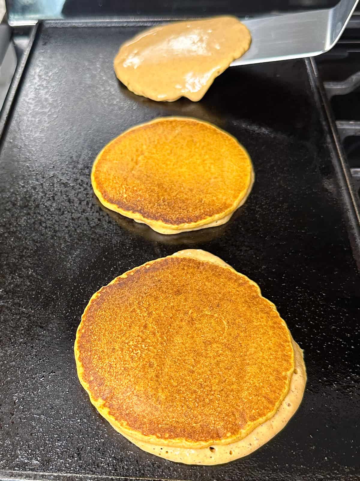 Three pumpkin pancakes on a griddle. The last pancakes is being flipped by a metal spatula.