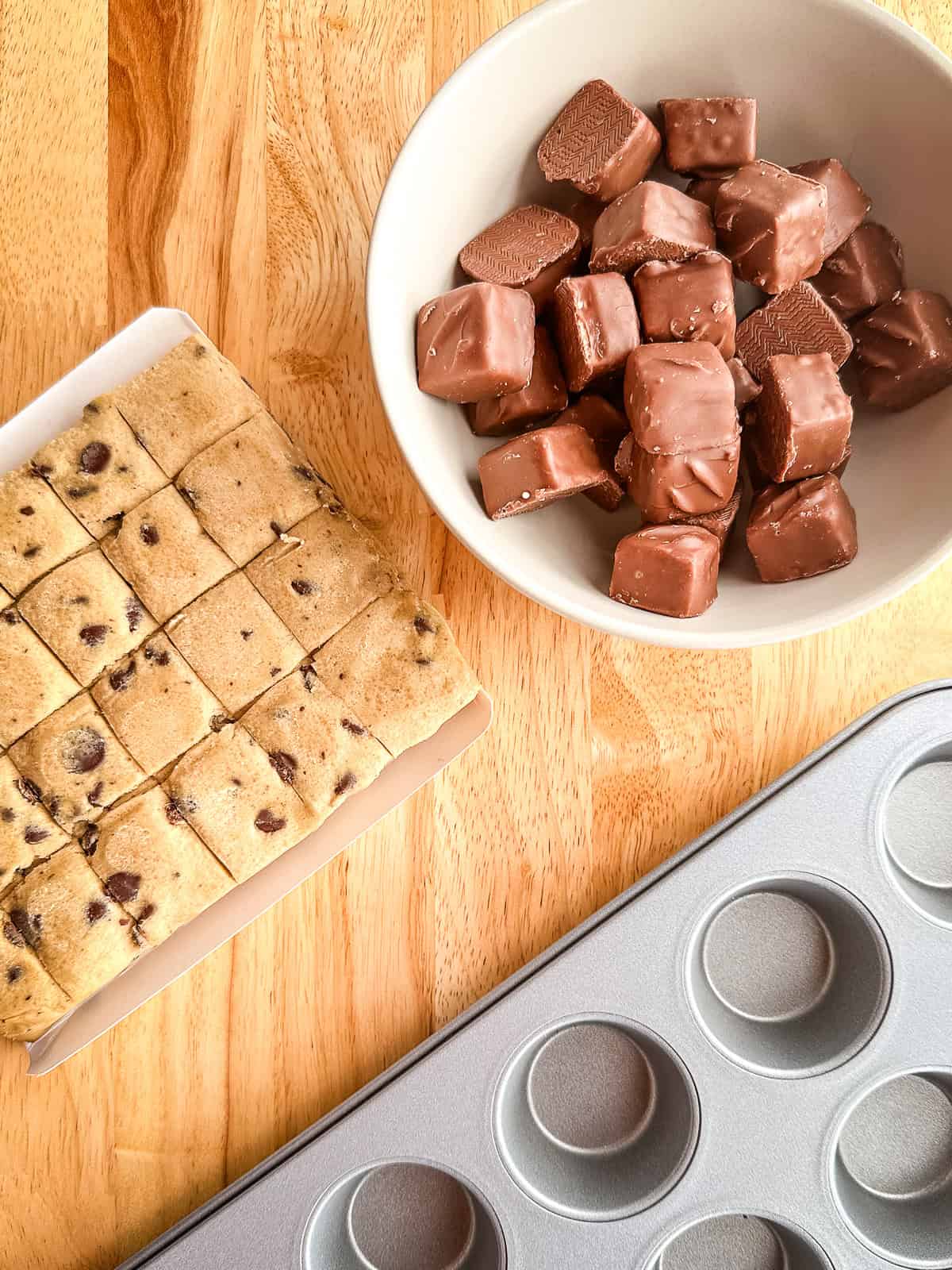 Ingredients for snickers cookie cups on the counter. A package of chocolate chip cookie dough and unwrapped Snickers mini candy bars. A mini muffin pan sits off to the side.