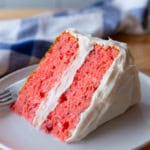 Strawberry cake slice on a white plate. The cake is frosted with cream cheese frosting.