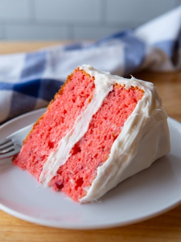 Strawberry cake slice on a white plate. The cake is frosted with cream cheese frosting.