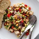 Chopped salad with tomatoes, cucumbers, olives, parsley, chicken, feta, chickpeas, and red onions on a plate with a fork, spoon and two slices of bread.