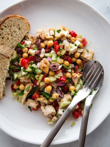 Chopped salad with tomatoes, cucumbers, olives, parsley, chicken, feta, chickpeas, and red onions on a plate with a fork, spoon and two slices of bread.
