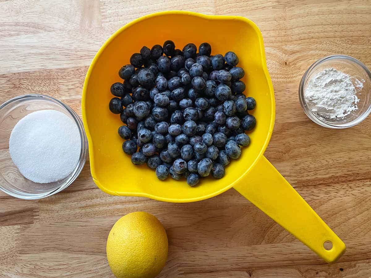 Ingredients for blueberry galette on the counter. The blueberries are in a yellow colander. The sugar and cornstarch are in separate glass bowls. And a lemon sits on the counter.