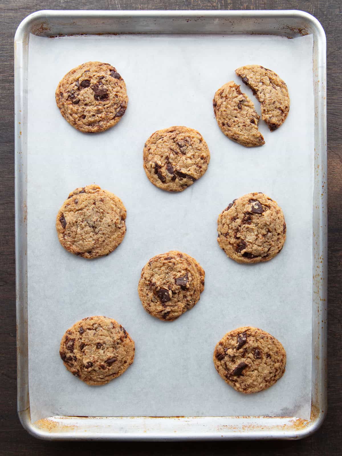 Eight chocolate chip cookies on a baking sheet. The cookie on the top right row is broken in half.